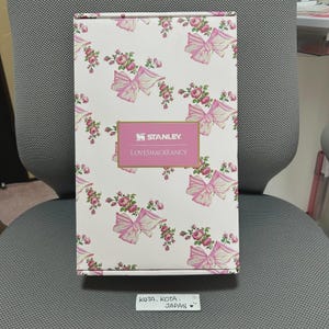 May include: A rectangular box with a white background, adorned with a repeating pattern of pink bows and floral arrangements. The box features the "STANLEY. LOVESHACKFANCY" logo in a pink rectangle. The box is sitting on a gray chair.