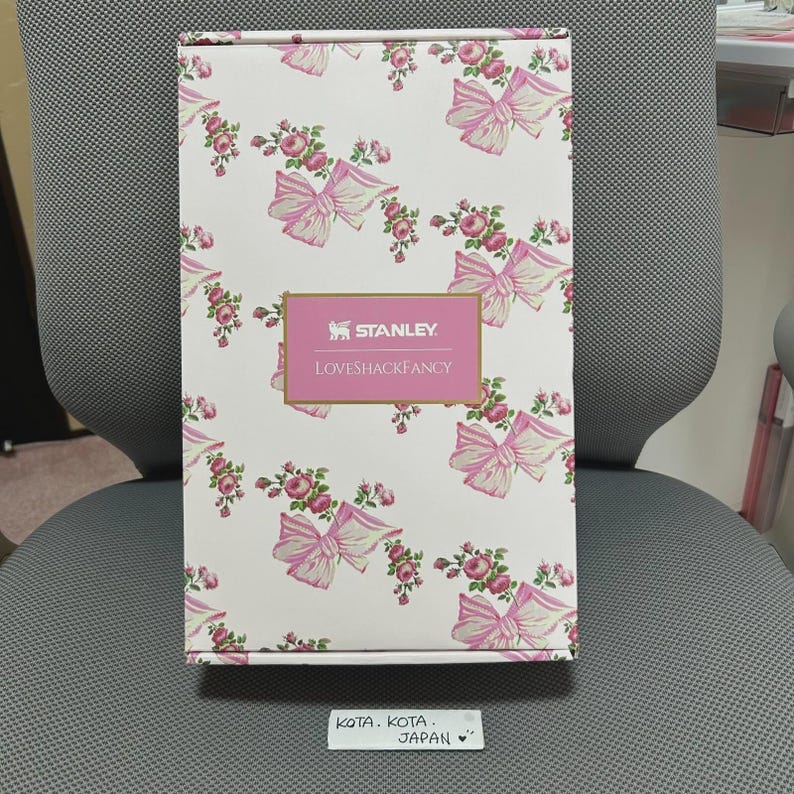 May include: A rectangular box with a white background, adorned with a repeating pattern of pink bows and floral arrangements. The box features the "STANLEY. LOVESHACKFANCY" logo in a pink rectangle. The box is sitting on a gray chair.