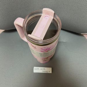 May include: A pink insulated tumbler with a handle and a clear lid. The tumbler features a floral design with pink flowers and green leaves. The lid has a sliding closure. The tumbler is sitting on a gray surface.