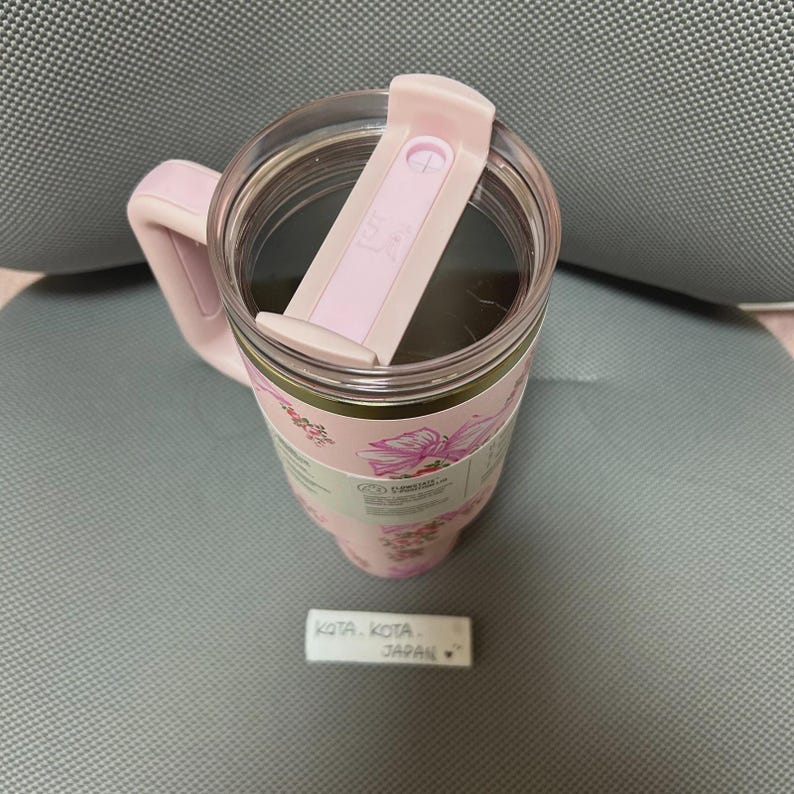 May include: A pink insulated tumbler with a handle and a clear lid. The tumbler features a floral design with pink flowers and green leaves. The lid has a sliding closure. The tumbler is sitting on a gray surface.