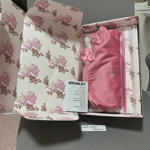 May include: An open box with a floral and bow pattern on the inside. Inside is a pink Stanley product, a pink carrying bag, and a pink straw. The box also contains a booklet with the Stanley logo.