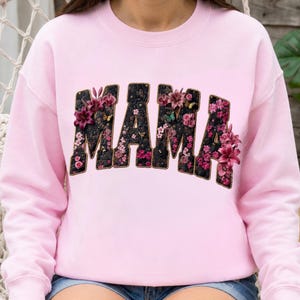 Pu&ograve; includere: Una felpa girocollo rosa chiaro con la parola MAMA in grandi lettere in stile blocco. Le lettere hanno uno sfondo nero testurizzato riempito con una variet&agrave; di motivi floreali rosa e viola, inclusi gigli e piccoli fiori.