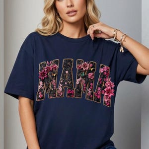 Pu&ograve; includere: Una t-shirt blu navy a maniche corte con la parola MAMA in grandi lettere stampatello. Le lettere sono riempite con un motivo floreale testurizzato nei toni del rosa, viola e verde, accentuato da piccole farfalle e bordi dorati.