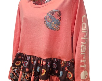Puede incluir: Camiseta de manga larga color coral con bolsillo de cachemira y dobladillo con volantes en capas. La manga lleva el nombre de la marca "Carhartt" en blanco. El volante inferior tiene un estampado floral y de cachemira.