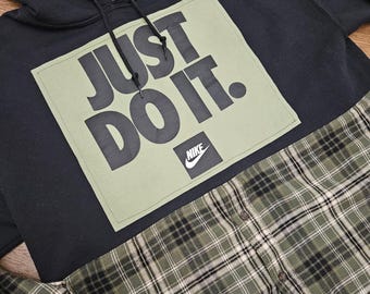 Puede incluir: Sudadera con capucha negra de Nike con el texto "JUST DO IT." en letras negras grandes sobre un cuadrado verde. Debajo, una camisa a cuadros verde, negra y blanca.