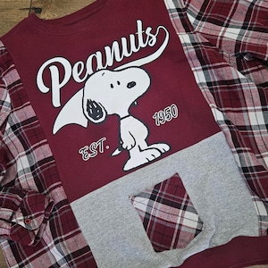 Pode incluir: Um moletom bordô com Snoopy de Peanuts, com a palavra "Peanuts" em escrita branca. O moletom tem detalhes em tecido cinza e xadrez, incluindo um bolso. O texto "EST. 1950" também é visível.