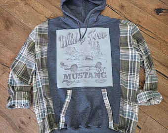 Puede incluir: Una sudadera con capucha gris con mangas a cuadros. La parte delantera presenta un gráfico azul claro con las palabras "Wild & Free" sobre un dibujo de un coche Mustang '65.