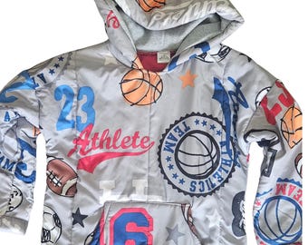 Puede incluir: Una sudadera con capucha gris con un estampado deportivo de balones de baloncesto, fútbol y fútbol americano. El diseño incluye el texto "Athlete", "Team Athletics", el número "23" en azul, el número "91" en rojo y varias estrellas.