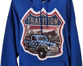 Puede incluir: Una sudadera con capucha azul real que presenta un gran parche gráfico en el pecho. El parche tiene forma de señal de carretera y muestra un camión antiguo con estrellas blancas, el texto TRADITION y el texto DRIVEN BY FREEDOM en letras rojas.