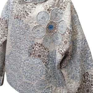 Könnte beinhalten: Ein gesteppter Kapuzenpullover in Blau-, Braun- und Cremetönen mit floralem Muster. Der Hoodie hat eine Vordertasche und ein dekoratives Blumendesign auf der Brust mit einem blauen Knopf in der Mitte. Das Kleidungsstück ist aus weichem, gestepptem Stoff gefertigt.