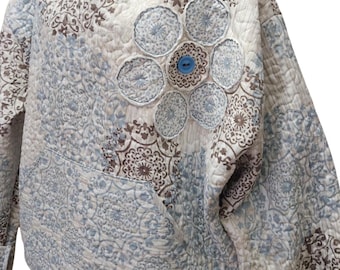 Puede incluir: Una sudadera con capucha acolchada en tonos azules, marrones y crema, con un estampado floral. La sudadera tiene un bolsillo delantero y un diseño floral decorativo en el pecho, con un botón azul en el centro. La prenda está hecha de tela acolchada suave.