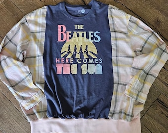 Puede incluir: Una camiseta de manga larga con cuello redondo y un panel central azul oscuro que presenta un gráfico de sol amarillo, las siluetas de The Beatles y el texto "THE BEATLES HERE COMES THE SUN". Las mangas y los laterales tienen un patrón de cuadros amarillo claro y rosa.