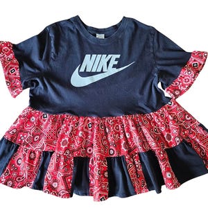 May include: Navy blue t-shirt dress with the Nike logo in white. The dress features red paisley print ruffle sleeves and a tiered skirt. A unique and stylish design.