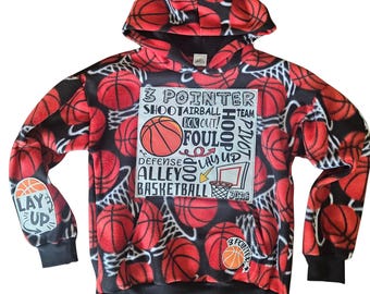 Puede incluir: Una sudadera con capucha negra con un estampado integral de balones de baloncesto naranjas y redes blancas. Un gráfico cuadrado central grande muestra el texto 3 POINTER, SHOOT, AIRBALL, BOX OUT, FOUL, HOOP, PIVOT, DEFENSE, LAY UP, ALLEY OOP, BASKETBALL, DUNK.