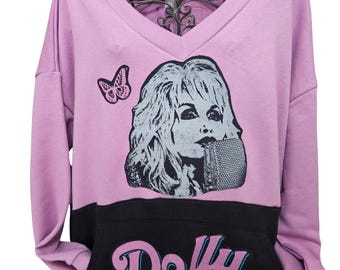 Puede incluir: Una sudadera lila con cuello en V con un gráfico en blanco y negro de una mujer con un micrófono. Una mariposa rosa está en la parte superior izquierda. La sección inferior es negra con el nombre "Dolly Parton" en una fuente estilizada.