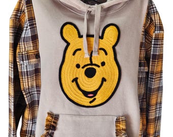 Puede incluir: Una sudadera con capucha beige con un gran parche amarillo con la cara de Winnie the Pooh. Las mangas son de tela a cuadros marrón, amarilla y blanca. La sudadera tiene un bolsillo delantero con un ribete con volantes y una capucha con cordón.
