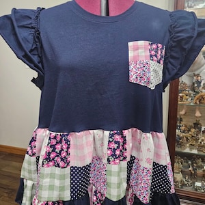May include: A navy blue short-sleeve shirt featuring ruffled sleeves and a chest pocket. The pocket and the lower section of the shirt are constructed from a patchwork of fabrics in pink gingham, white with black polka dots, and floral patterns.