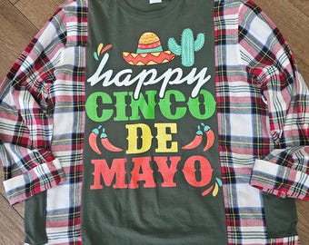 Puede incluir: Camiseta de manga larga verde oliva con un gráfico colorido de "Happy Cinco de Mayo". El diseño incluye un sombrero, un cactus y chiles. Las mangas son de un patrón de cuadros rojos, blancos y negros.