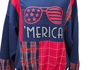 Puede incluir: Una sudadera azul marino con detalles rojos y parches a cuadros. La parte delantera presenta unas gafas de sol con estrellas y rayas, y la palabra 'MERICA' en plata. Las mangas también tienen detalles a cuadros.