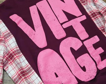 Puede incluir: Una sudadera granate con la palabra VINTAGE en letras rosas grandes y desgastadas en el pecho. La prenda tiene mangas largas de tela a cuadros blanca, roja y granate, que contrastan con el torso de color granate sólido.