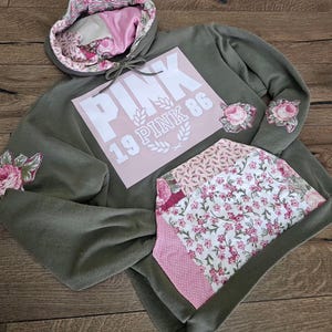 Puede incluir: Una sudadera con capucha verde oliva con un bolsillo y forro de capucha con estampado floral rosa y blanco. La parte delantera presenta un cuadrado rosa con la palabra "PINK" y los números "19 PINK 86" en blanco. Parches florales adornan las mangas.