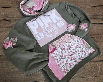 Puede incluir: Una sudadera con capucha verde oliva con un bolsillo y forro de capucha con estampado floral rosa y blanco. La parte delantera presenta un cuadrado rosa con la palabra "PINK" y los números "19 PINK 86" en blanco. Parches florales adornan las mangas.
