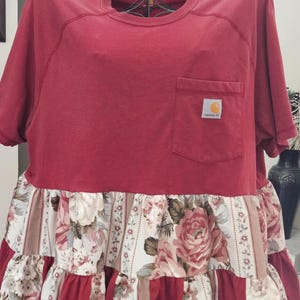 Op de afbeelding: Rode t-shirtjurk met een bloemenrok. De top heeft een zak met een Carhartt-logo. De rok heeft een mix van bloemen- en gestreepte stof in tinten rood, crème en bruin. De jurk heeft korte mouwen.