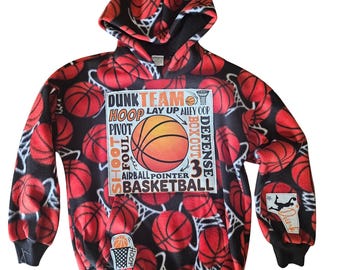 Puede incluir: Una sudadera con capucha de forro polar negro con un estampado integral de balones de baloncesto naranjas y redes blancas. Un gráfico central rectangular muestra terminología de baloncesto, incluyendo DUNK, TEAM, HOOP, LAY UP, ALLEY OOP, PIVOT, BOX OUT, DEFENSE, SHOOT, FOUL, AIRBALL, POINTER y BASKETBALL.