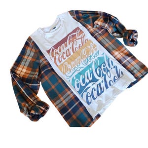 May include: A white long-sleeved shirt with a Coca-Cola logo design in red, orange, yellow, teal, and blue. The shirt is layered with a plaid flannel shirt in shades of orange, blue, and brown.