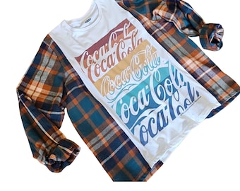Puede incluir: Una camiseta blanca de manga larga con el logotipo de Coca-Cola en rojo, naranja, amarillo, verde azulado y azul. Está superpuesta con una camisa de franela a cuadros en tonos naranja, azul y marrón.