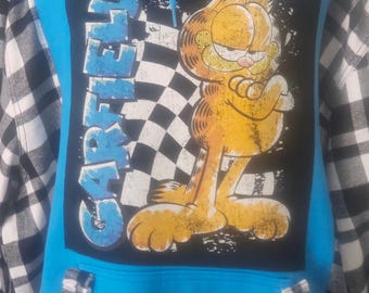 Puede incluir: Sudadera con capucha azul con un gráfico de Garfield sobre un fondo negro con la palabra "GARFIELD" en azul. Las mangas son de cuadros blancos y negros. La sudadera tiene un cordón y un bolsillo delantero.