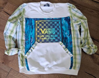 Puede incluir: Sudadera blanca con diseño tie-dye azul y logotipo de Vans. Las mangas son de una camisa a cuadros verde y amarilla. La sudadera tiene un bolsillo delantero y el texto "Vans Off The Wall Since 1966".