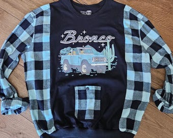 Puede incluir: Sudadera negra con mangas y bolsillo a cuadros azules y negros. La parte delantera presenta un gráfico de camioneta Bronco azul con la palabra "Bronco" en escritura blanca. También se incluye un gráfico de cactus.