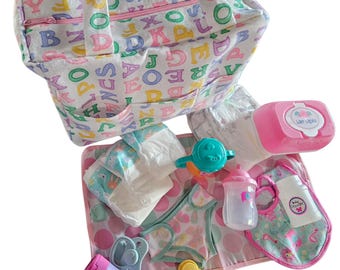 Puede incluir: Un bolso de tela con estampado de alfabeto colorido que contiene un juego de accesorios para muñecas, incluyendo un cambiador, pañales, una caja de toallitas húmedas rosa, un biberón, un chupete, loción, polvos y un sonajero amarillo.