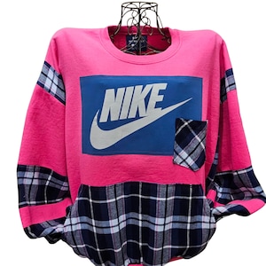 Op de afbeelding: Een felroze sweater met ronde hals en een grote blauwe rechthoekige patch met het witte Nike-logo en swoosh. Het kledingstuk heeft een borstzak met een marineblauw, wit en lichtblauw ruitpatroon en bijpassende geruite stoffen panelen op de mouwen en het onderste voorpand.