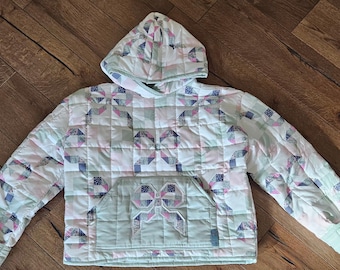 Puede incluir: Una sudadera con capucha acolchada que presenta un patrón de patchwork de cuadrados de tela blancos, verde salvia y florales en rosa y azul. La prenda incluye un bolsillo canguro delantero grande con un detalle acolchado en forma de lazo.