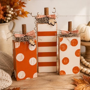 May include: Three rectangular wooden blocks shaped like pumpkins, featuring orange and white paint. Two blocks have white polka dots on orange, and one has orange stripes on white. Each has a brown stem, a gingham ribbon, and dried moss.