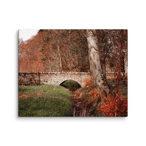 Pode incluir: Impressão em tela de uma ponte de pedra com um único arco sobre um pequeno riacho. A paisagem circundante inclui árvores com folhas castanho-avermelhadas e relva verde, criando uma cena de outono.