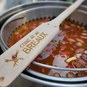 May include: A wooden paddle with the text "COME AT ME BREAUX" and a crawfish illustration is positioned over a pot of food. The paddle is light-colored, and the text and crawfish are etched into the wood. The pot is stainless steel and filled with a stew-like dish.