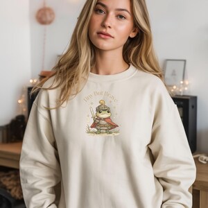 Op de afbeelding: Een crèmekleurige sweatshirt met een grillige illustratie van een kikker gekleed als ridder, compleet met helm, cape en zwaard. De tekst "Tiny But Brave" staat boven de kikker gedrukt. De sweatshirt heeft lange mouwen.