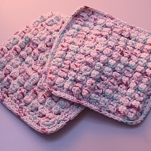 May include: Two square crochet dishcloths featuring a textured bobble stitch pattern. The yarn is a light pink base color with small, speckled flecks of blue, purple, and red throughout the fabric. Each cloth has a simple single crochet border.