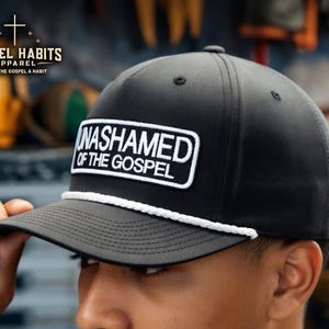 May include: Black baseball cap with a white rope detail and a rectangular patch that reads "UNASHAMED OF THE GOSPEL" in white lettering. The cap has a curved brim and is being held by a person.