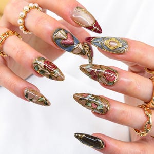 May include: Close-up of elaborately decorated long fingernails. Each nail showcases a unique stained-glass-style design with gold detailing. The designs include floral patterns, hearts, and geometric shapes in various colours, including red, blue, and green.