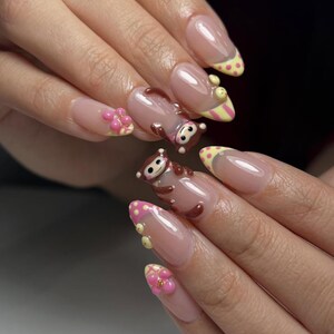 Könnte beinhalten: Lange, ovale Acrylnägel mit klarem Grund und French-Spitzen. Die Spitzen sind mit gelben und rosa Punkten und Streifen verziert. Einige Nägel haben 3D-Affen- und Blumen-Designs für einen verspielten Look.