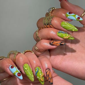 May include: Long, pointed fingernails with detailed nail art. Designs include ladybirds on blue and green backgrounds, green leaves with water droplets, caterpillars, and butterfly wings. Gold rings adorn the fingers.