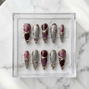 May include: A set of ten long, pointed artificial nails in a clear plastic case. The nails feature a silver metallic design with burgundy and grey accents. The nails are arranged in two rows of five.