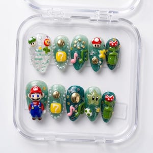 May include: A set of ten press-on nails featuring a Super Mario theme. The nails are in a clear plastic case and showcase various designs, including Mario, question blocks, mushrooms, and stars. The nails are primarily green, white, and red.