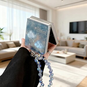 以下が含まれることがあります： 青と白の銀河のデザインの透明な携帯電話ケースで、雲の形をした2つの装飾が特徴です。ケースには、虹色のビーズとシルバーのハードウェアが付いたビーズのリストストラップが付いています。ケースのテキストは「sweetest of my path, beautiful dreams」と書かれています。