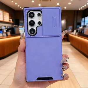Pode incluir: Uma capa de telefone lilás com uma tampa deslizante para a câmera. A capa tem formato retangular com borda preta e recorte para as lentes da câmera do telefone. A tampa da câmera é de um tom lilás mais claro.