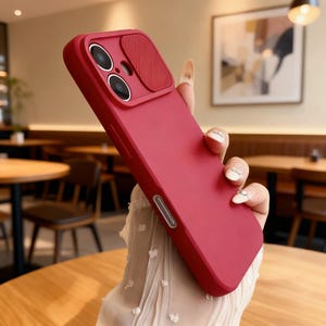 Puede incluir: Una funda roja para teléfono con una cubierta para la cámara. La funda tiene un acabado liso y una sección elevada alrededor de las lentes de la cámara. La funda se sostiene en ángulo, mostrando su diseño y color.